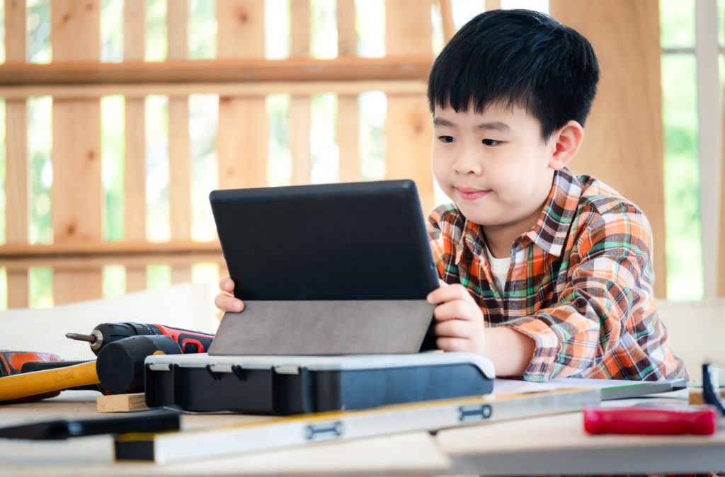 Young Asia boy wearing a shirt and sitting at the table at home and see the tablet. Many equipment tools (hammer, screwdriver, saw) on the table. Carpenter and education concept. Concentrate on tablet